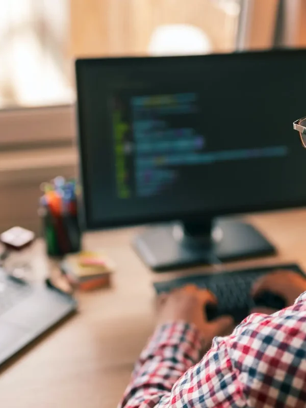 Person at a desk with a laptop and monitor displaying code, in a well-lit home or office setting.